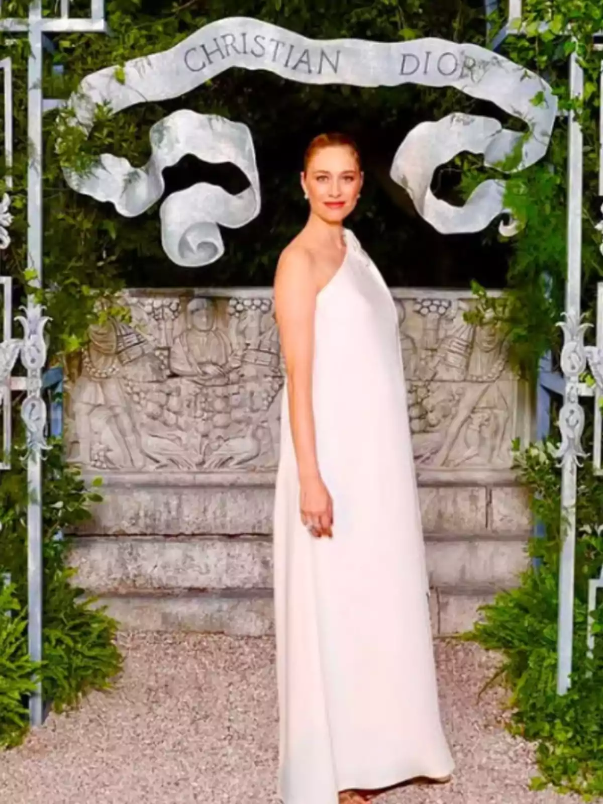 Woman in a long white dress posing at an outdoor event under a decorative arch with the inscription Christian Dior, surrounded by greenery Woman in a long white dress posing at an outdoor event under a decorative arch with the inscription Christian Dior, surrounded by greenery