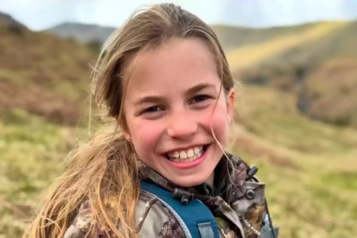 Una niña sonriente con el cabello rubio suelto en un entorno natural con colinas verdes al fondo. Una niña sonriente con el cabello rubio suelto en un entorno natural con colinas verdes al fondo.