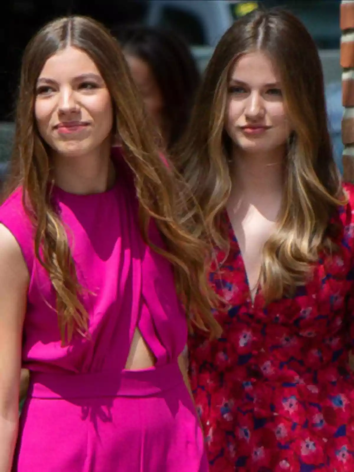Dos chicas jóvenes con el cabello largo y suelto posan al aire libre, una lleva un vestido fucsia y la otra un vestido rojo con estampado floral Dos chicas jóvenes con el cabello largo y suelto posan al aire libre, una lleva un vestido fucsia y la otra un vestido rojo con estampado floral