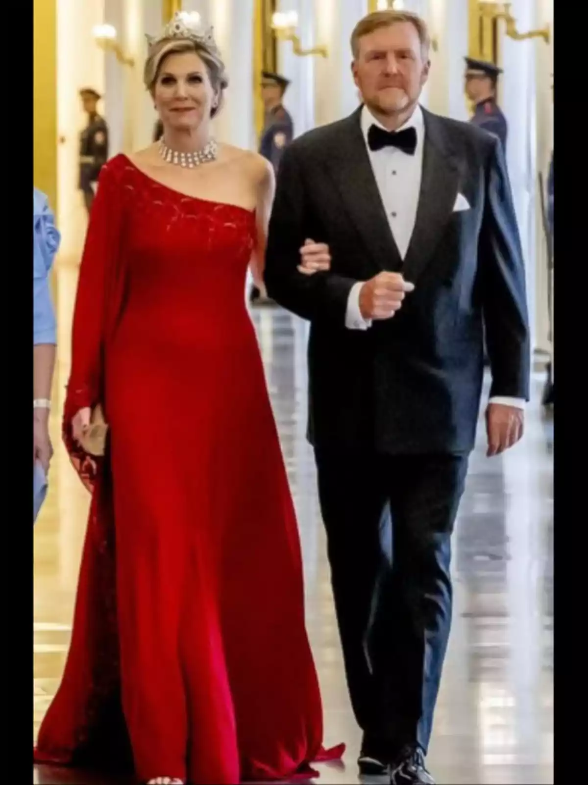 Elegantly dressed couple walks down a luxurious hallway; she wears a long red dress and a tiara while he is dressed in a black tuxedo.