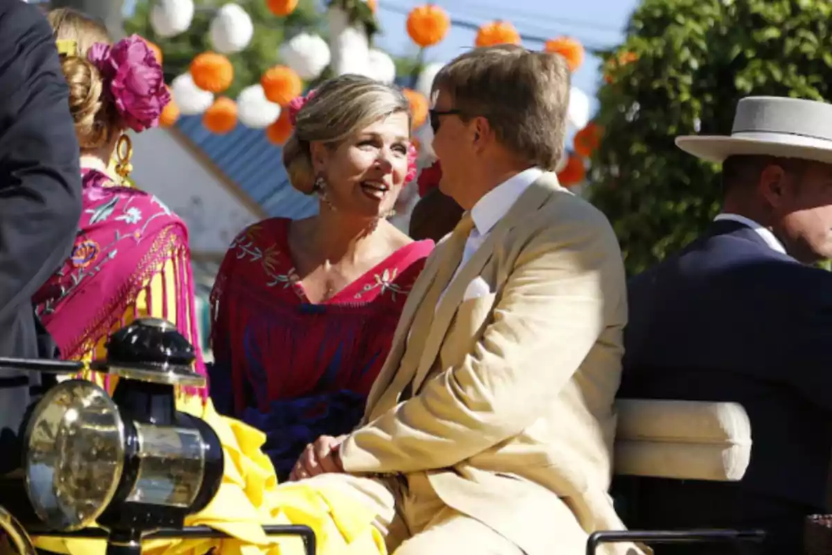 A couple dressed in traditional costumes chats happily while riding in a carriage decorated with paper flowers during an outdoor celebration.