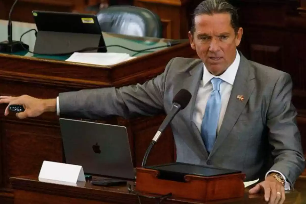 A man in a gray suit and blue tie is standing at a podium with a microphone, next to a laptop in a conference room.