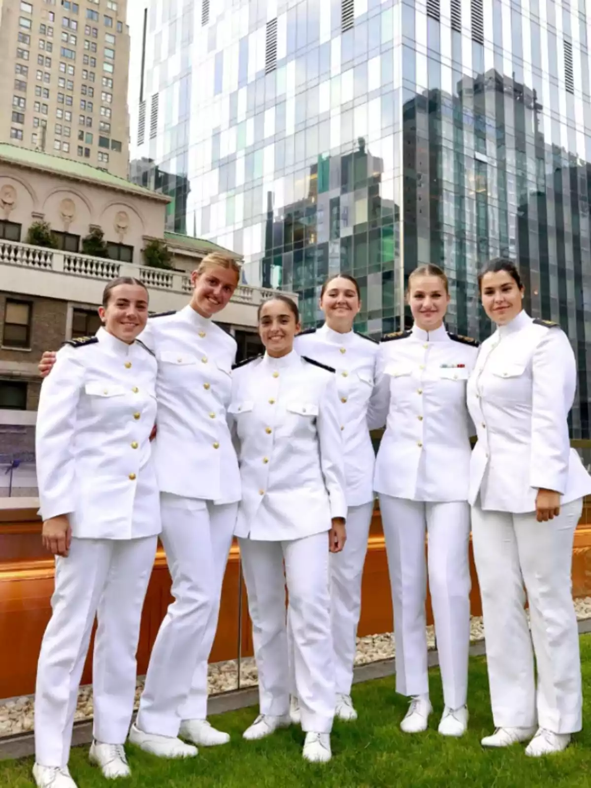 Six women dressed in formal white uniforms pose smiling outdoors in front of modern and old buildings. Six women dressed in formal white uniforms pose smiling outdoors in front of modern and old buildings.