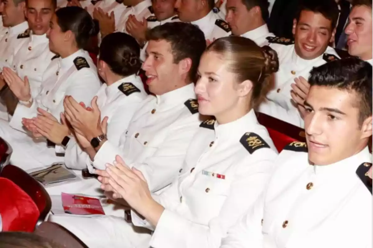 A group of cadets dressed in white uniforms sitting in an auditorium, applauding and smiling during a formal event.