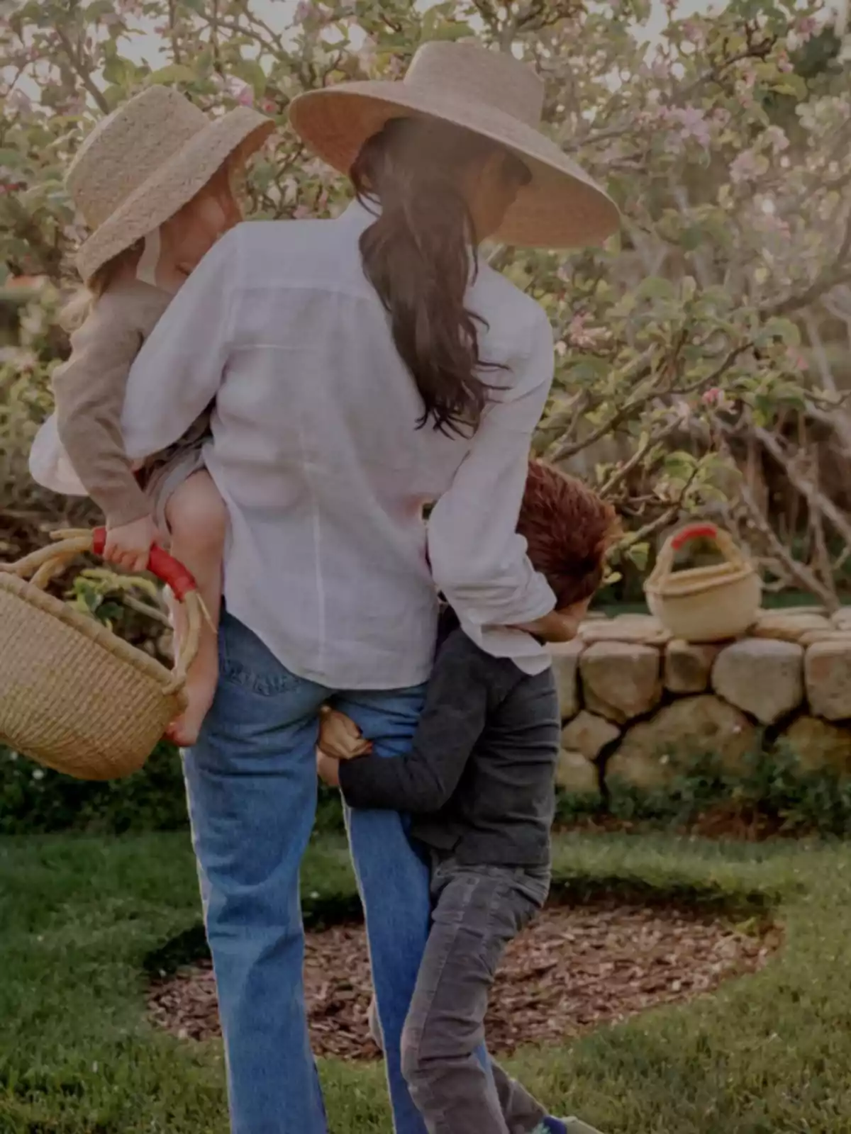 Una mujer de espaldas con sombrero sostiene a una niña pequeña mientras un niño la abraza de la pierna en un jardín con flores y césped Una mujer de espaldas con sombrero sostiene a una niña pequeña mientras un niño la abraza de la pierna en un jardín con flores y césped