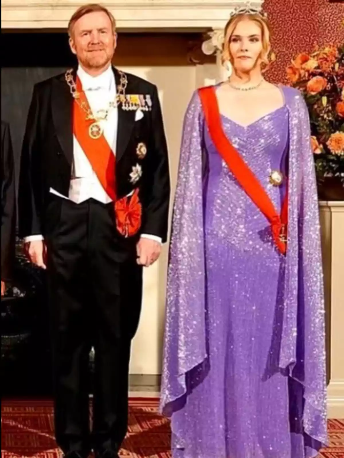 Pareja vestida de gala, el hombre lleva esmoquin negro con banda roja y medallas, la mujer lleva un vestido largo morado brillante, tiara y banda roja, ambos posan en un entorno elegante con flores al fondo Pareja vestida de gala, el hombre lleva esmoquin negro con banda roja y medallas, la mujer lleva un vestido largo morado brillante, tiara y banda roja, ambos posan en un entorno elegante con flores al fondo