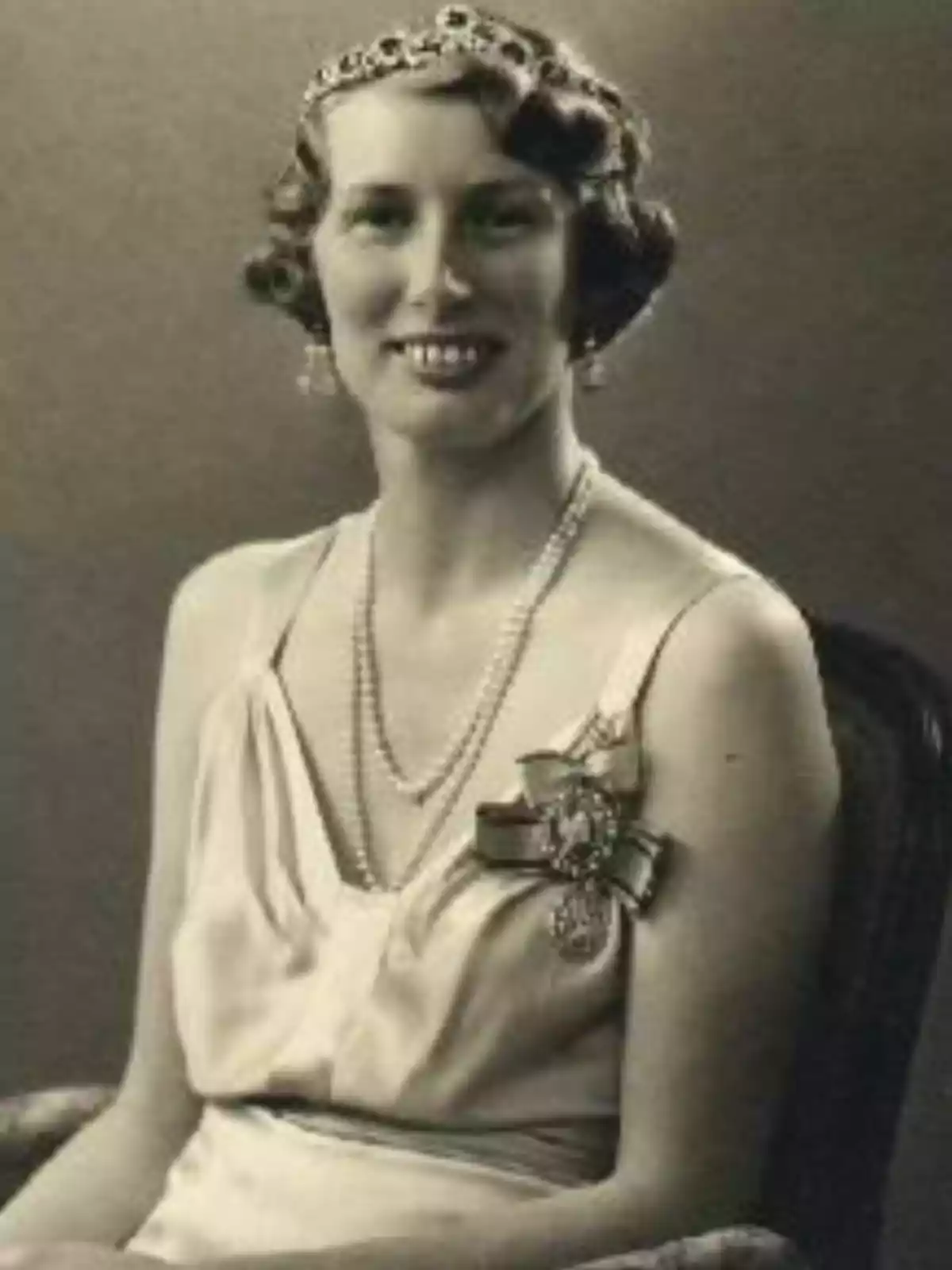 Mujer joven con vestido elegante, tiara, collar de perlas y broche, sentada y sonriendo en una fotografía antigua en blanco y negro Mujer joven con vestido elegante, tiara, collar de perlas y broche, sentada y sonriendo en una fotografía antigua en blanco y negro