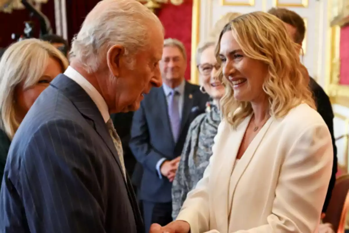 An older man with gray hair and a striped suit greets a smiling blonde woman with wavy hair and a white blazer in an elegant room with red walls and gold details, while other people watch in the background. An older man with gray hair and a striped suit greets a smiling blonde woman with wavy hair and a white blazer in an elegant room with red walls and gold details, while other people watch in the background.
