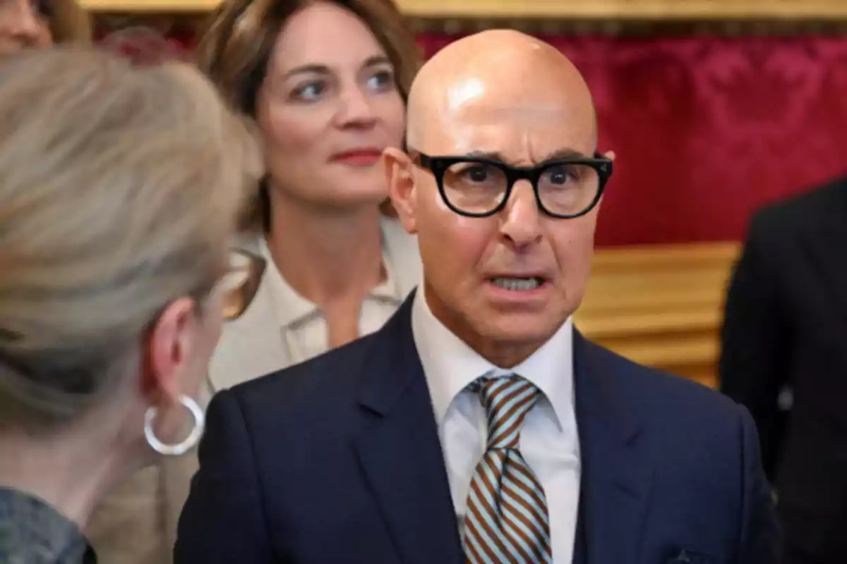 Bald man with glasses and a blue suit talking to a woman with blonde hair tied back while another woman watches in the background in an elegant setting with red walls. Bald man with glasses and a blue suit talking to a woman with blonde hair tied back while another woman watches in the background in an elegant setting with red walls.