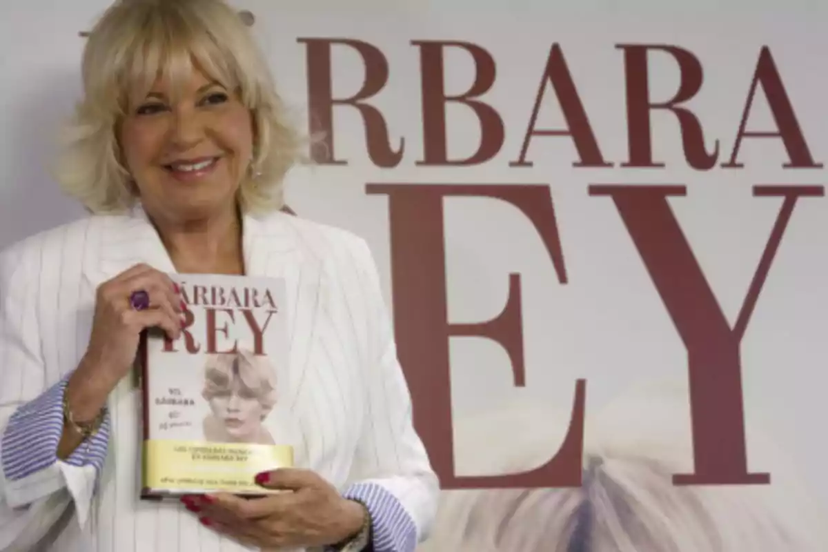 Mujer rubia de cabello corto y sonrisa vistiendo un saco blanco a rayas sostiene un libro con su imagen en la portada frente a un cartel grande con letras que dicen Bárbara Rey Mujer rubia de cabello corto y sonrisa vistiendo un saco blanco a rayas sostiene un libro con su imagen en la portada frente a un cartel grande con letras que dicen Bárbara Rey