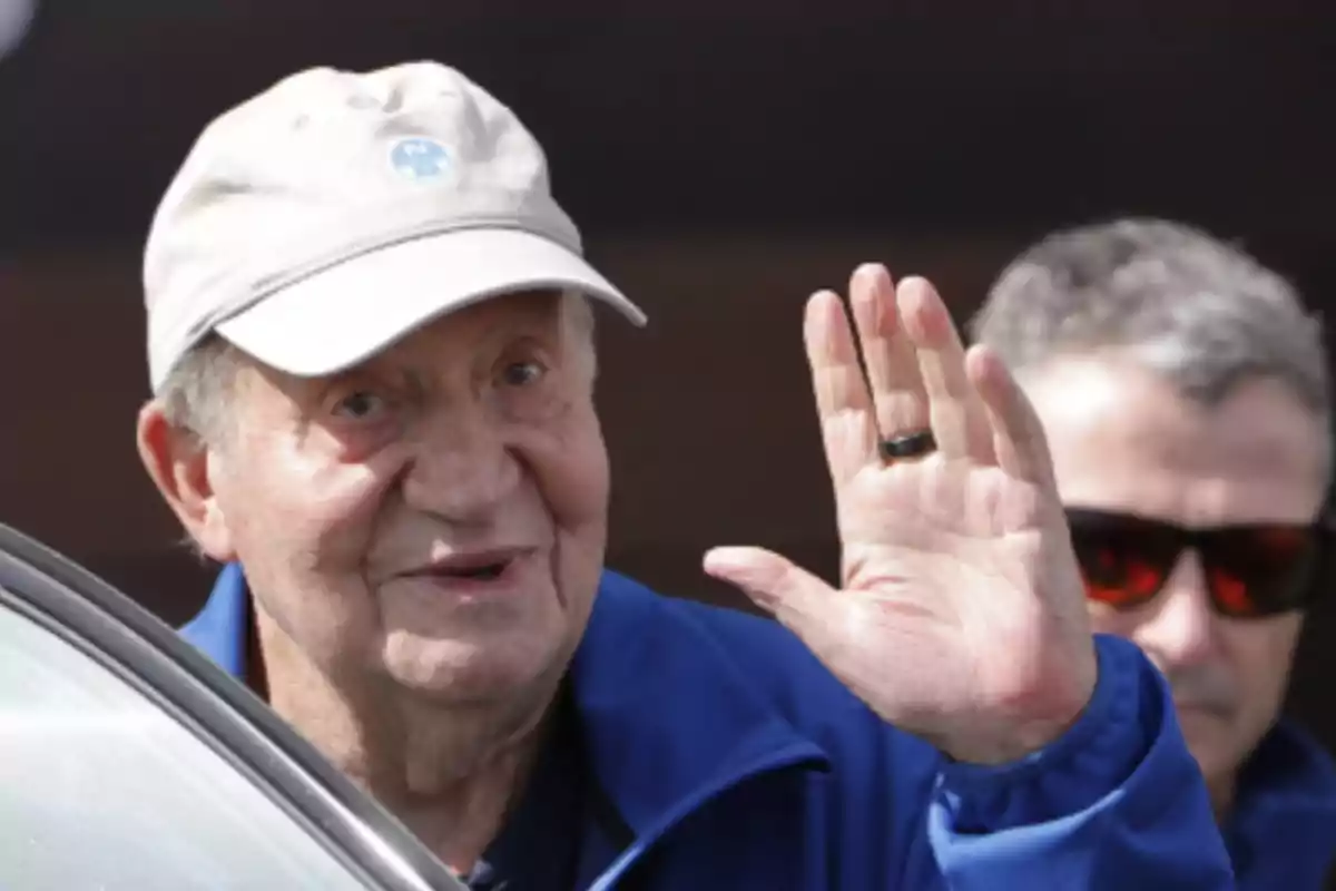 Hombre mayor con gorra clara y chaqueta azul saludando con la mano levantada mientras otra persona con gafas oscuras está al fondo Hombre mayor con gorra clara y chaqueta azul saludando con la mano levantada mientras otra persona con gafas oscuras está al fondo