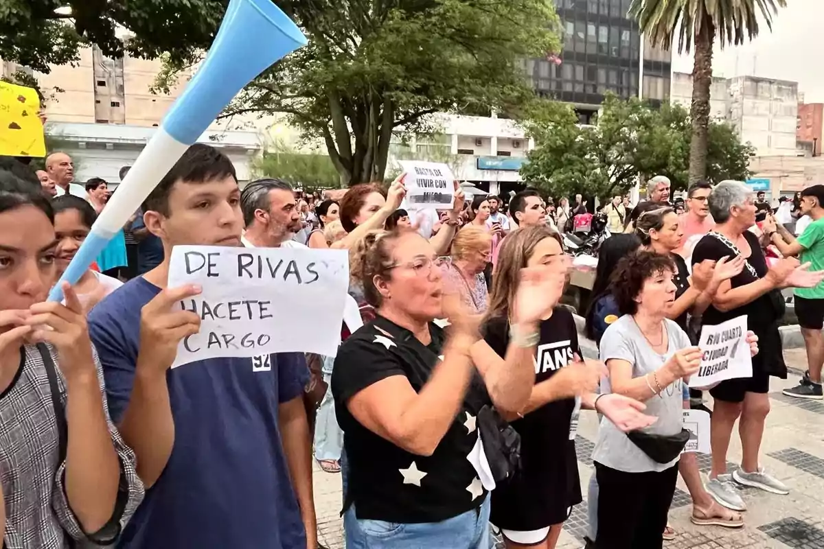 Personas manifestándose en la calle con carteles y una vuvuzela, rodeadas de árboles y edificios. Personas manifestándose en la calle con carteles y una vuvuzela, rodeadas de árboles y edificios.