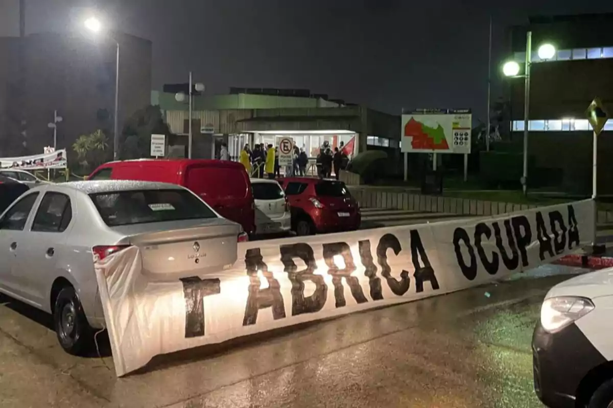 Several cars parked in front of a factory at night with a large tarp that says FACTORY OCCUPIED and people gathered at the entrance of the building. Several cars parked in front of a factory at night with a large tarp that says FACTORY OCCUPIED and people gathered at the entrance of the building.