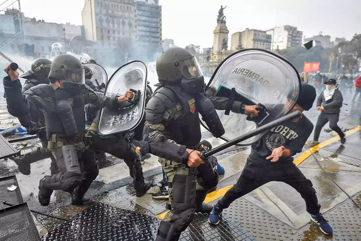 Policías antidisturbios con equipo de protección y escudos enfrentan a manifestantes en una calle urbana.