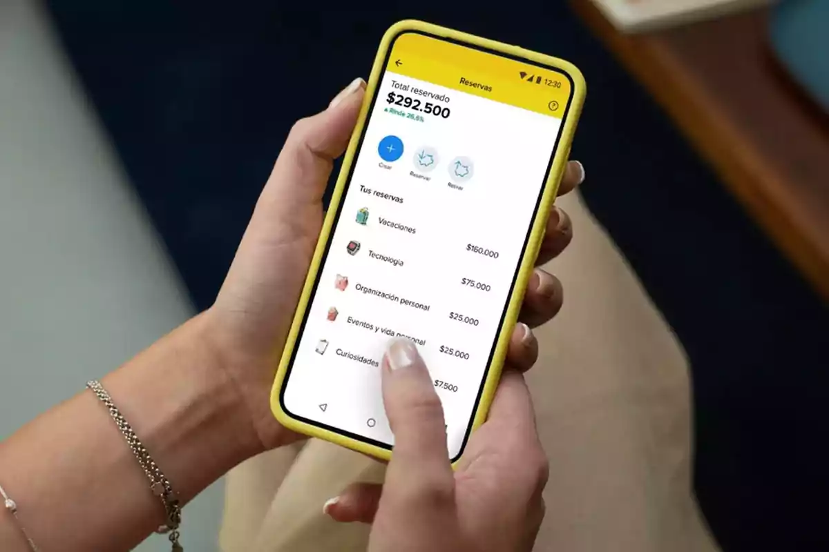 Persona sosteniendo un teléfono móvil con una aplicación de finanzas que muestra diferentes categorías de ahorro y el monto total reservado Persona sosteniendo un teléfono móvil con una aplicación de finanzas que muestra diferentes categorías de ahorro y el monto total reservado