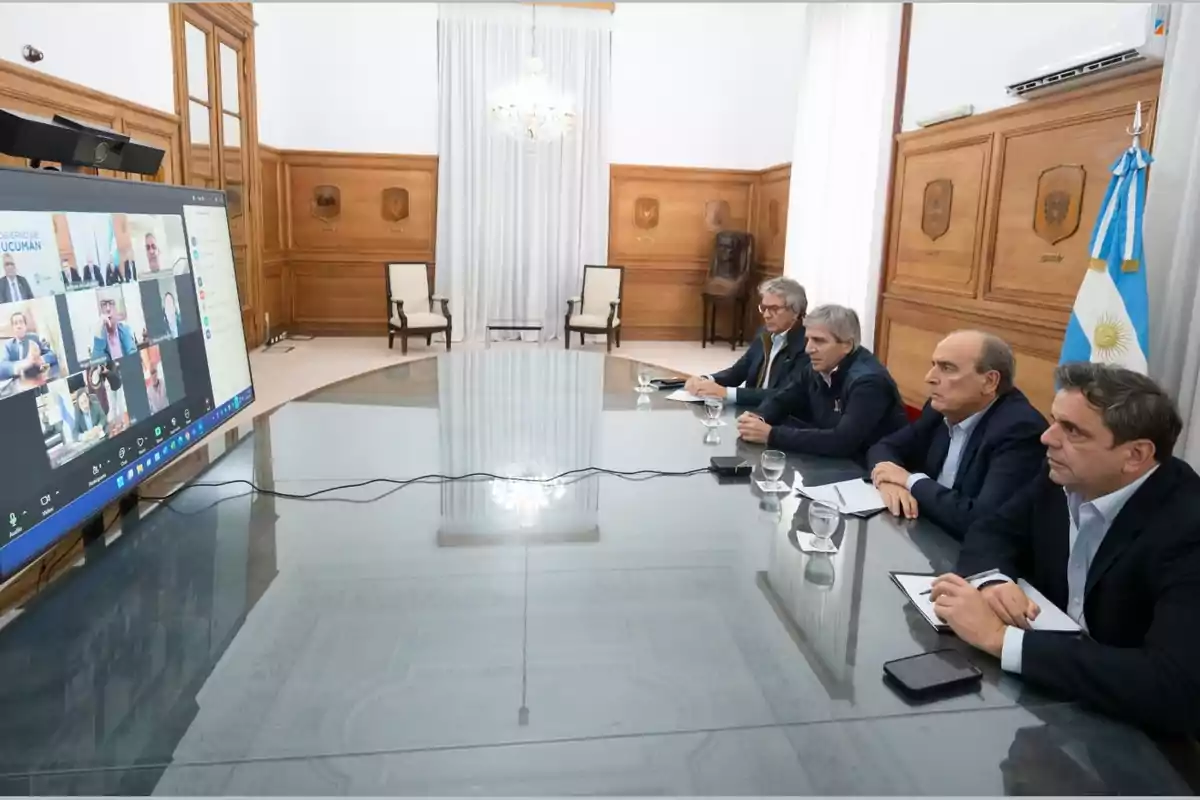 Four men seated at a conference table participate in a group video call in an official room with an Argentine flag in the background. Four men seated at a conference table participate in a group video call in an official room with an Argentine flag in the background.