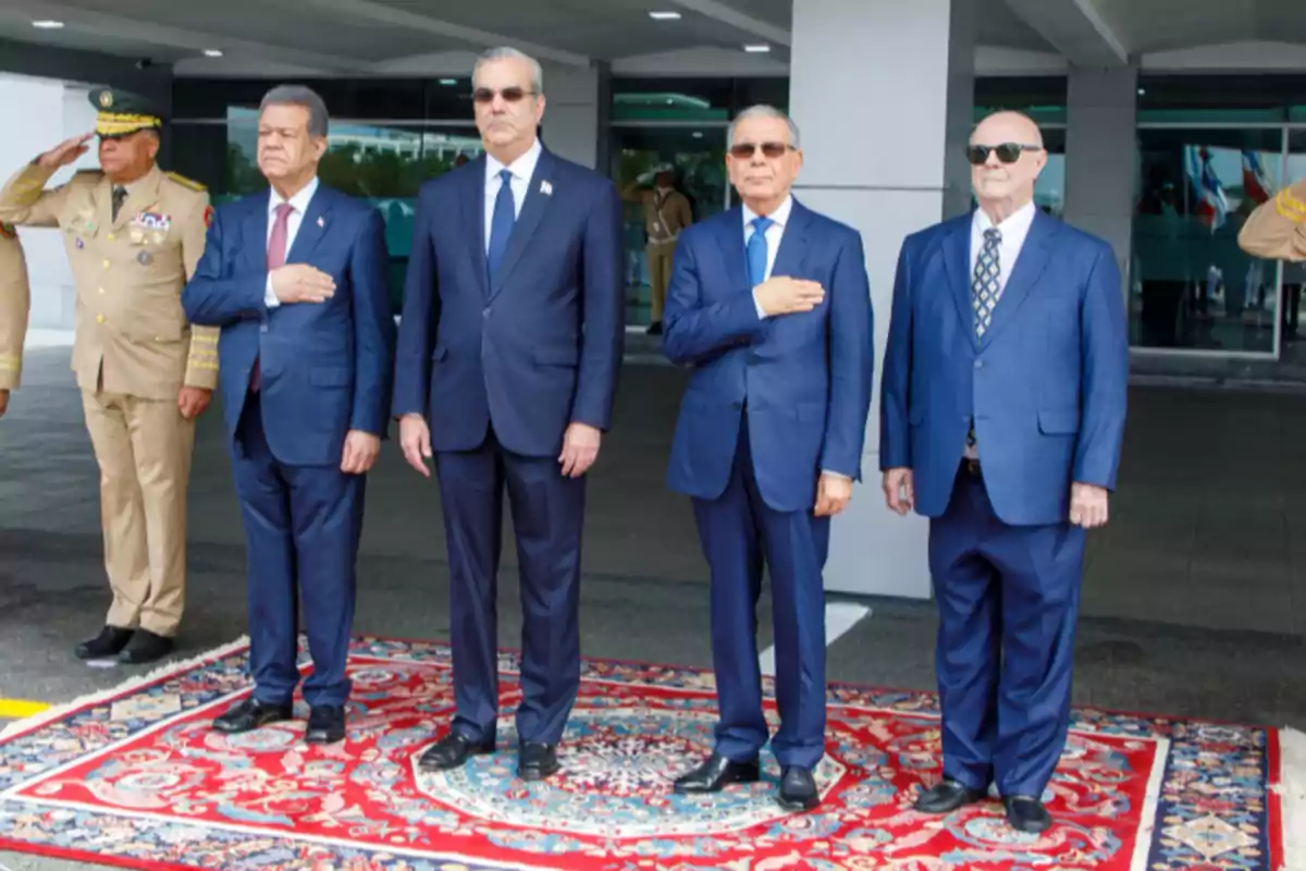 Un grupo de hombres en trajes formales se encuentra de pie sobre una alfombra decorativa en un evento oficial al aire libre, con un militar saludando al fondo.