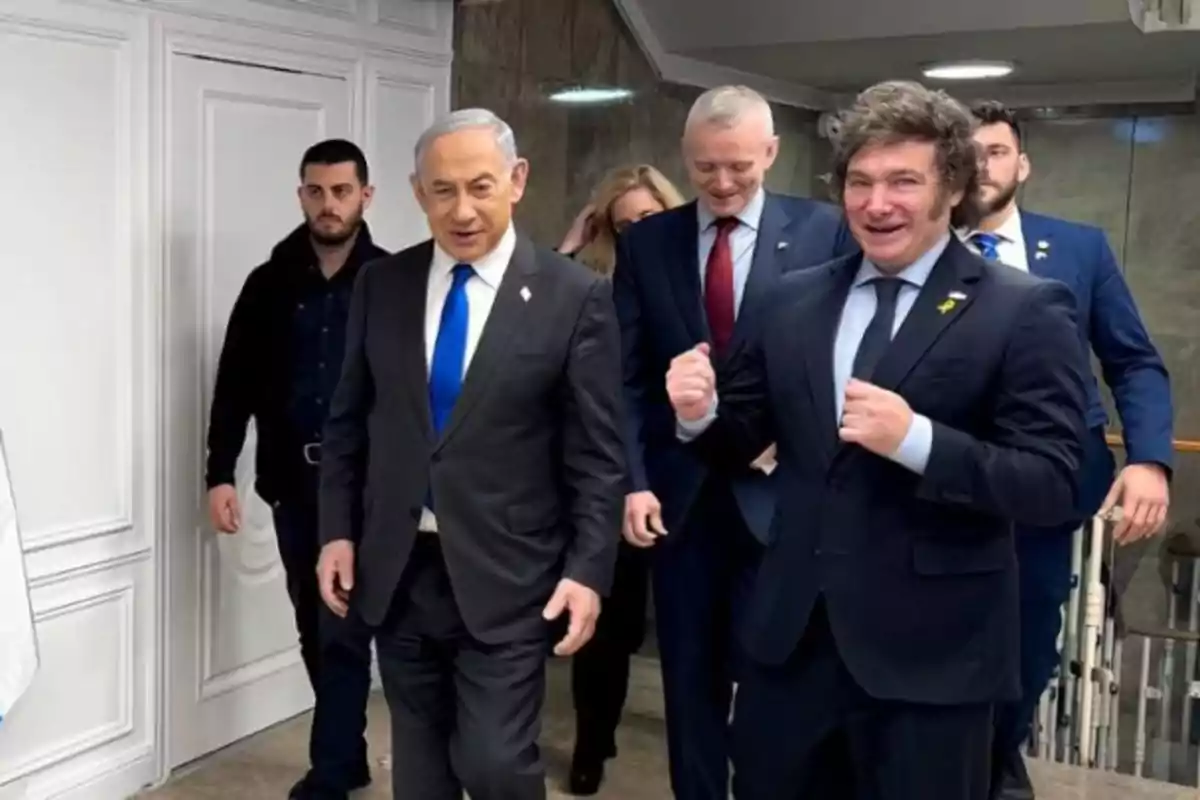 Un grupo de personas vestidas de traje camina por un pasillo mientras dos de ellos al frente sonríen y parecen conversar animadamente Un grupo de personas vestidas de traje camina por un pasillo mientras dos de ellos al frente sonríen y parecen conversar animadamente