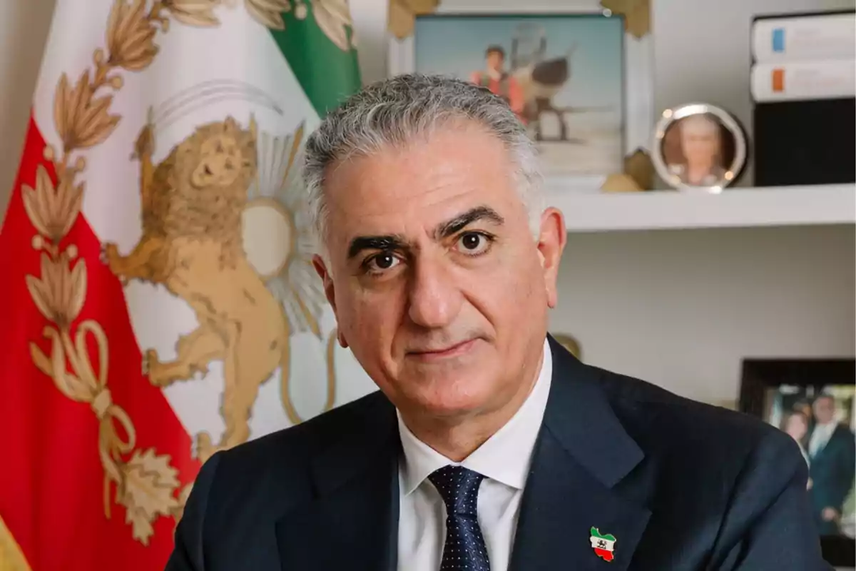 Hombre de cabello canoso con traje oscuro y corbata, posando frente a una bandera con un león dorado y fondo de estantería con fotos y libros. Hombre de cabello canoso con traje oscuro y corbata, posando frente a una bandera con un león dorado y fondo de estantería con fotos y libros.