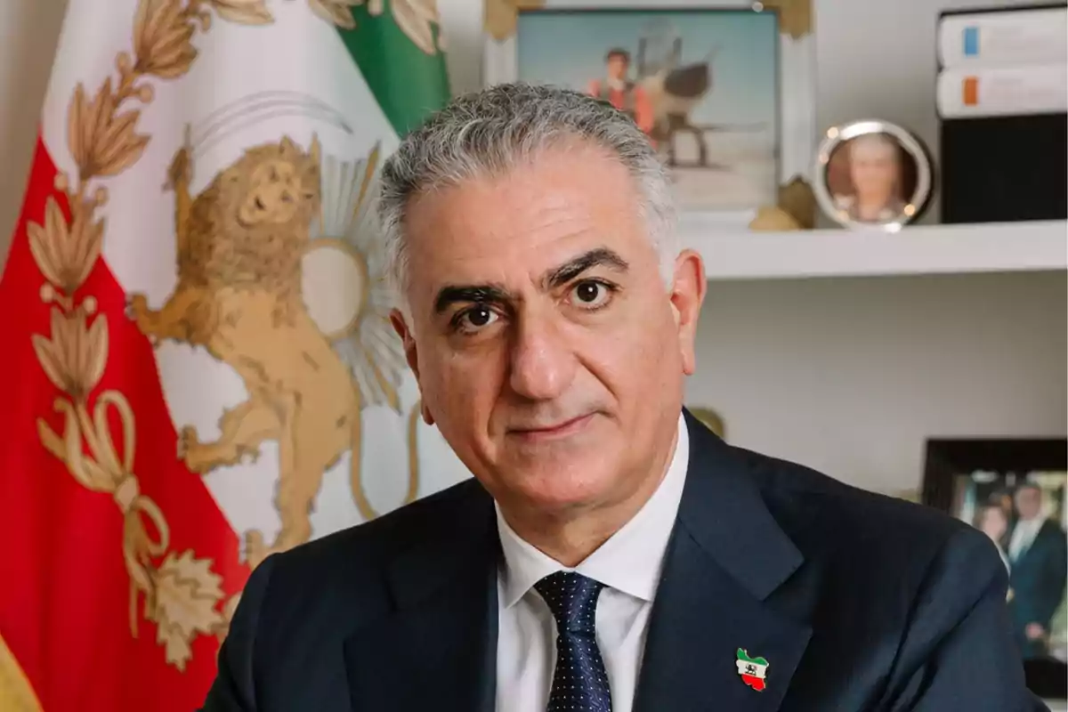 Hombre de cabello canoso con traje oscuro y corbata, posando frente a una bandera con un león y decoraciones doradas, con estantería y fotos familiares al fondo