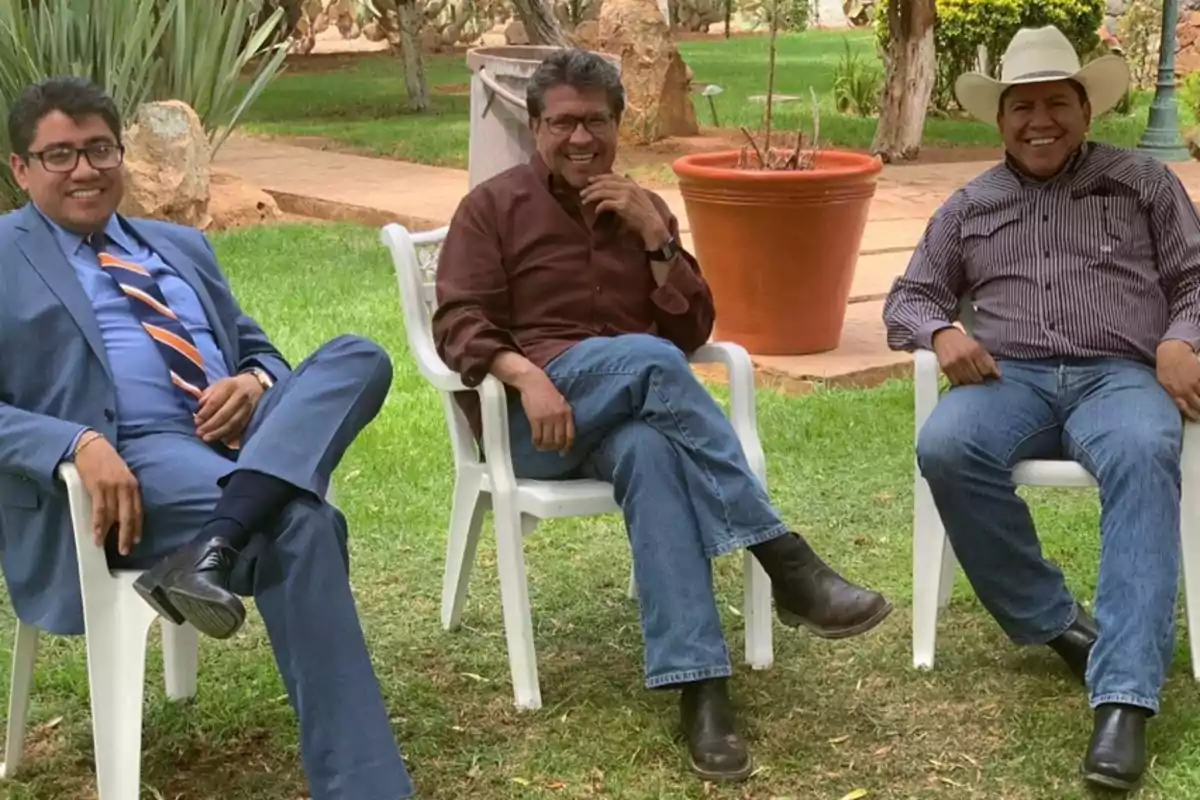 Tres hombres sentados en sillas de plástico blancas en un jardín, conversando y sonriendo, uno de ellos lleva traje azul, otro camisa marrón y el tercero camisa a rayas y sombrero blanco. Tres hombres sentados en sillas de plástico blancas en un jardín, conversando y sonriendo, uno de ellos lleva traje azul, otro camisa marrón y el tercero camisa a rayas y sombrero blanco.