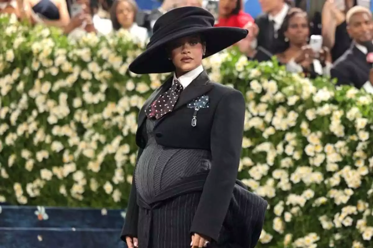 A person in an elegant black suit and large hat poses in front of a background of white and yellow flowers while people watch in the background. A person in an elegant black suit and large hat poses in front of a background of white and yellow flowers while people watch in the background.