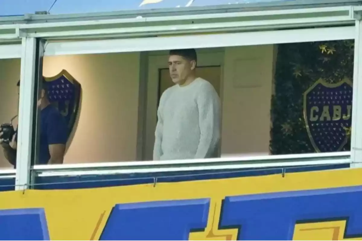 A man in a white sweater is standing in a stadium box, looking out, with soccer team crests on the wall behind him. A man in a white sweater is standing in a stadium box, looking out, with soccer team crests on the wall behind him.