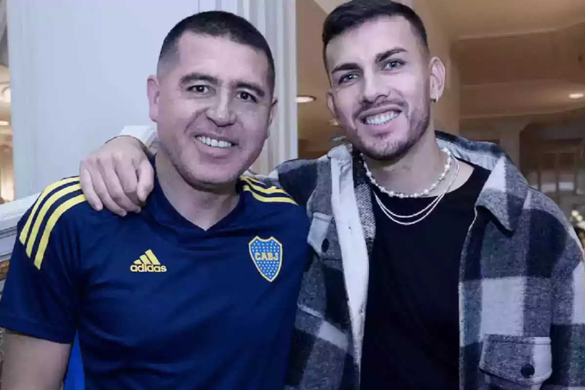 Two men smile and pose together; one is wearing a blue sports jersey with the Boca Juniors crest, and the other is dressed in a plaid jersey and necklaces. Two men smile and pose together; one is wearing a blue sports jersey with the Boca Juniors crest, and the other is dressed in a plaid jersey and necklaces.