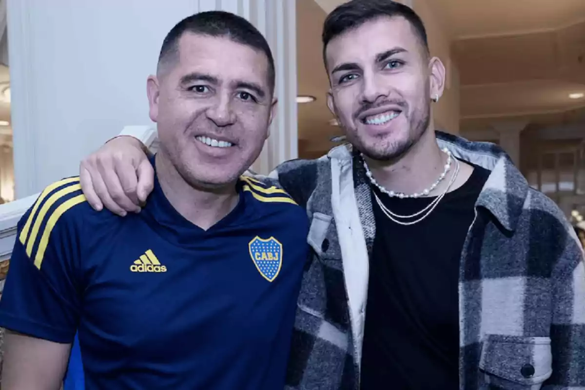 Dos hombres sonríen y posan juntos, uno lleva una camiseta deportiva azul con detalles amarillos y el otro viste una camisa a cuadros en tonos grises y negros Dos hombres sonríen y posan juntos, uno lleva una camiseta deportiva azul con detalles amarillos y el otro viste una camisa a cuadros en tonos grises y negros