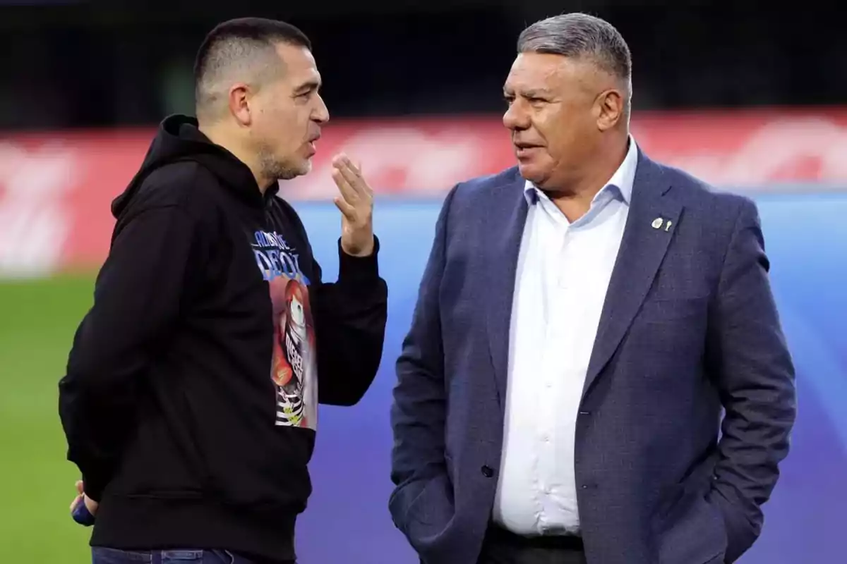 Dos hombres conversando en un campo de fútbol, uno con sudadera negra y el otro con traje azul.