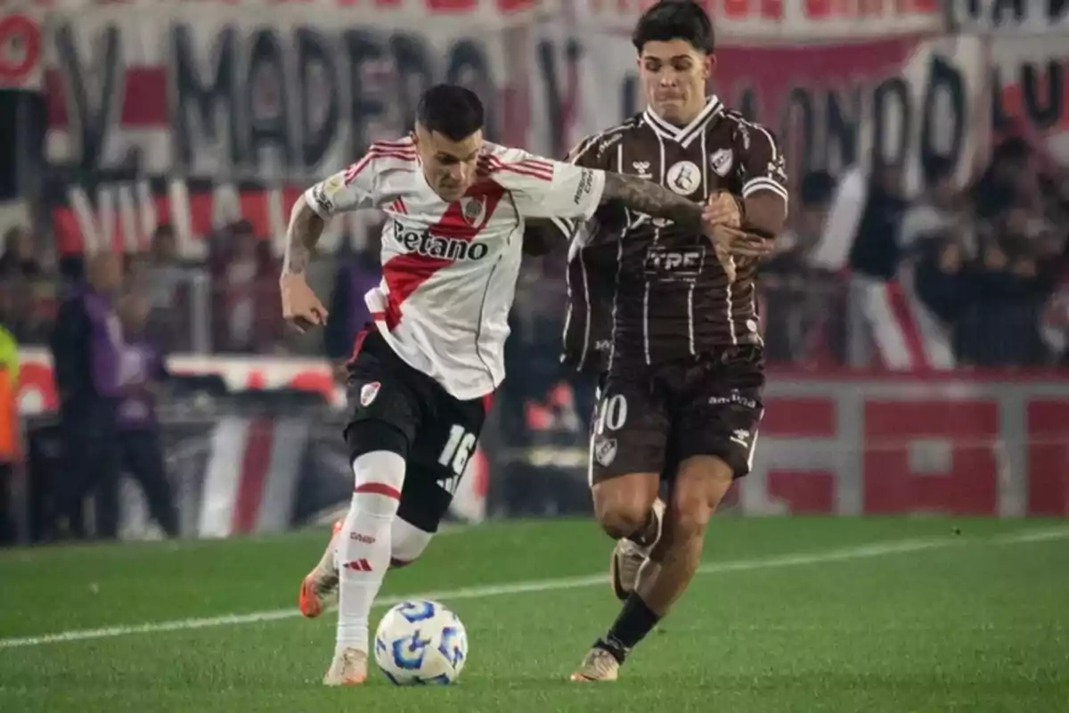 Two players compete for the ball in a match, one in a white and red uniform and the other in a brown uniform, while the crowd watches from the stands. Two players compete for the ball in a match, one in a white and red uniform and the other in a brown uniform, while the crowd watches from the stands.