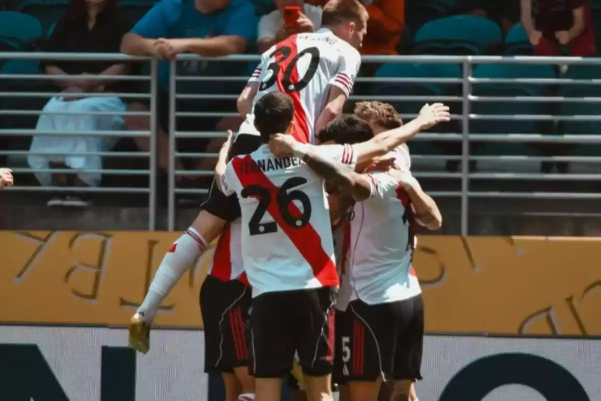 Soccer players wearing white uniforms with a red stripe celebrate a goal by hugging on the field in front of the stands.