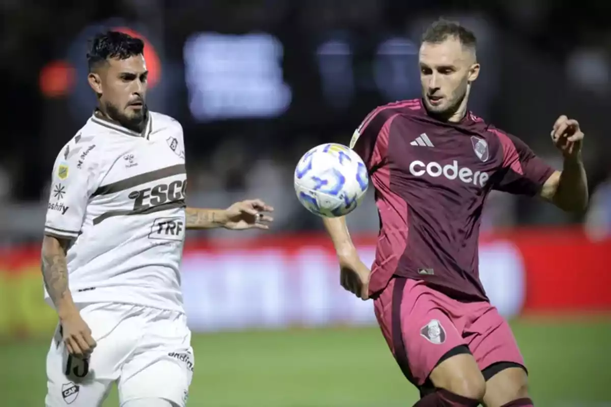 Dos jugadores de fútbol en acción durante un partido, uno con uniforme blanco y el otro con uniforme granate, ambos concentrados en el balón. Dos jugadores de fútbol en acción durante un partido, uno con uniforme blanco y el otro con uniforme granate, ambos concentrados en el balón.
