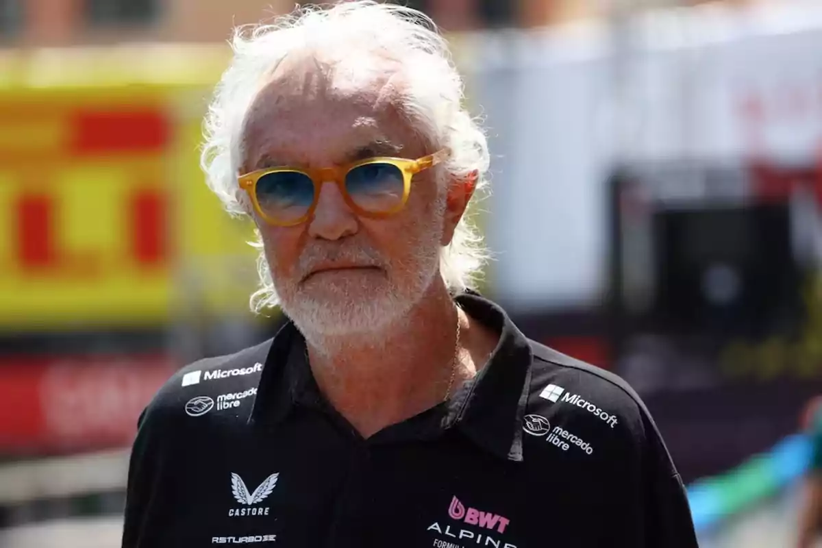 A man with white hair and a beard is wearing orange sunglasses and a black jersey with various brand logos. A man with white hair and a beard is wearing orange sunglasses and a black jersey with various brand logos.