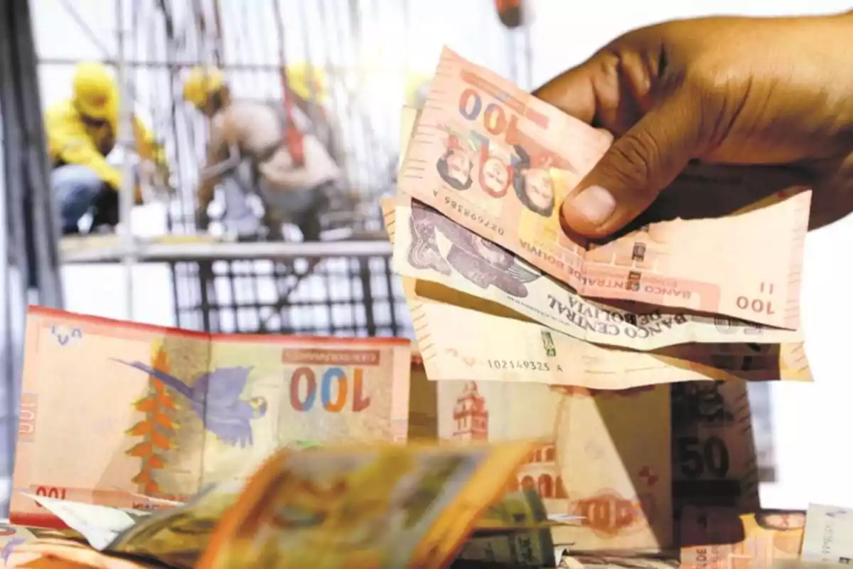 Una mano sosteniendo billetes de bolivianos mientras al fondo se ven trabajadores de la construcción.
