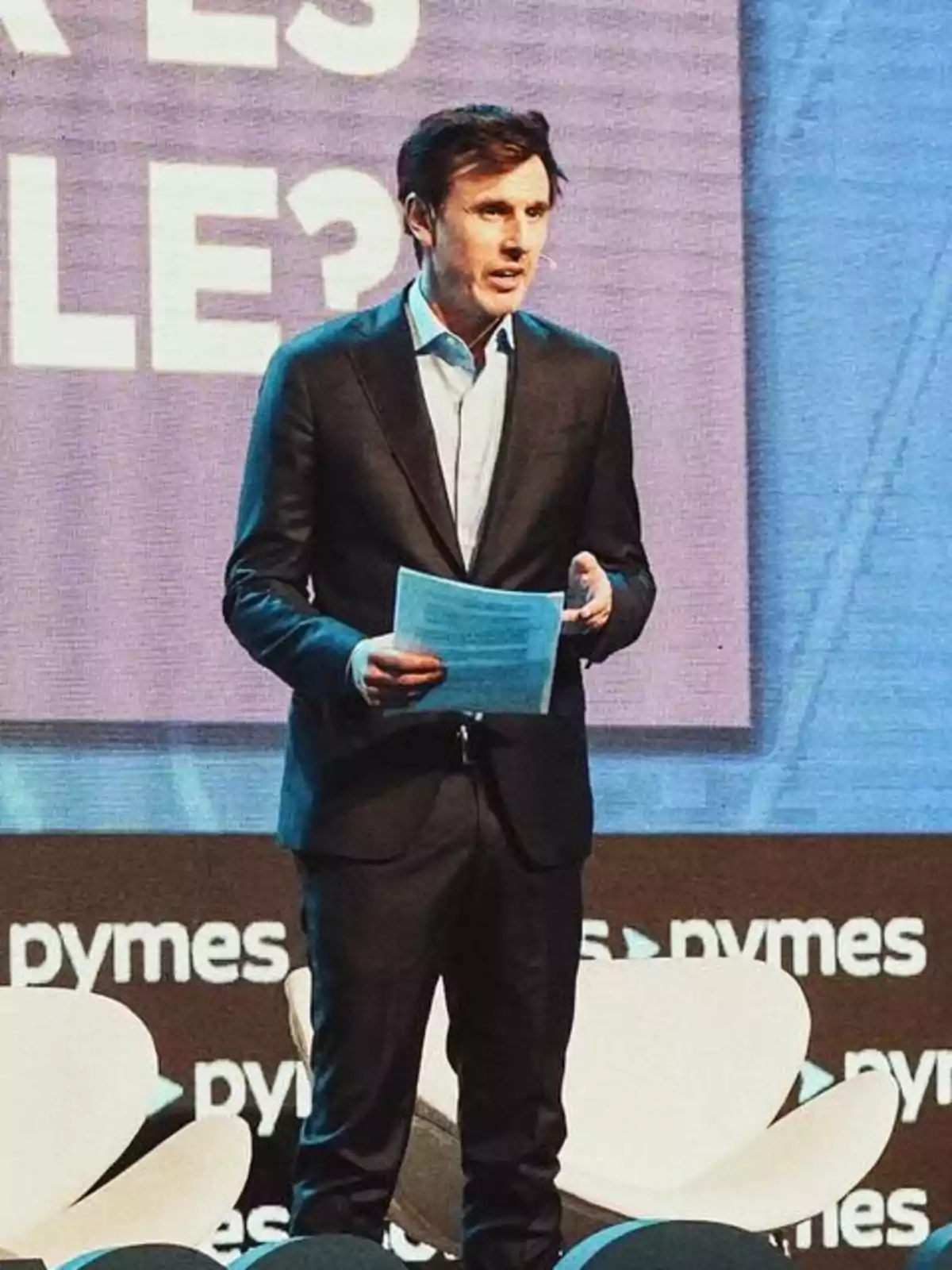 Hombre de traje oscuro hablando en un escenario con fondo de letras y sillas blancas Hombre de traje oscuro hablando en un escenario con fondo de letras y sillas blancas
