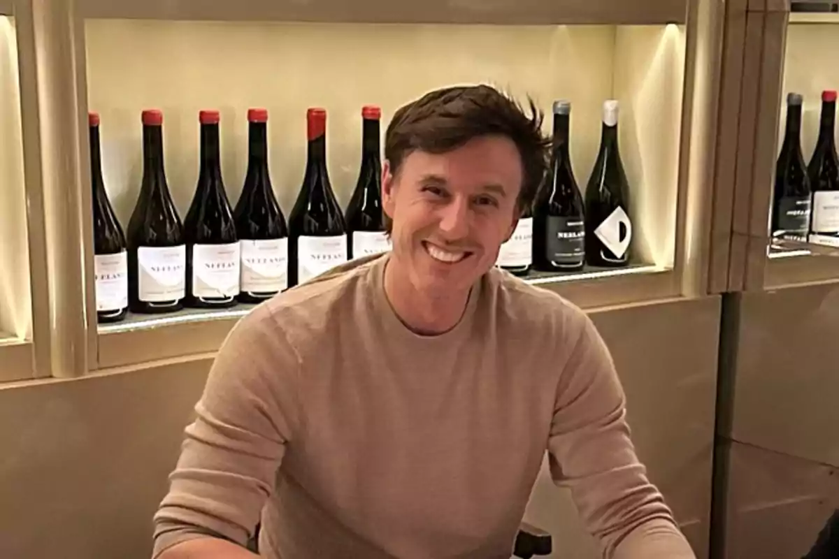 Hombre sonriente sentado frente a una estantería con botellas de vino