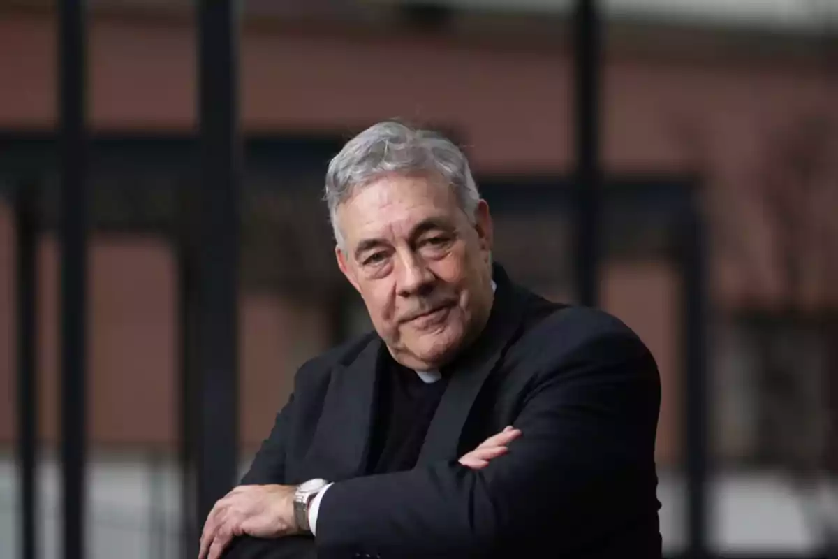 An older man with gray hair and a serious expression wears a dark suit and poses with his arms crossed in front of a blurred background.