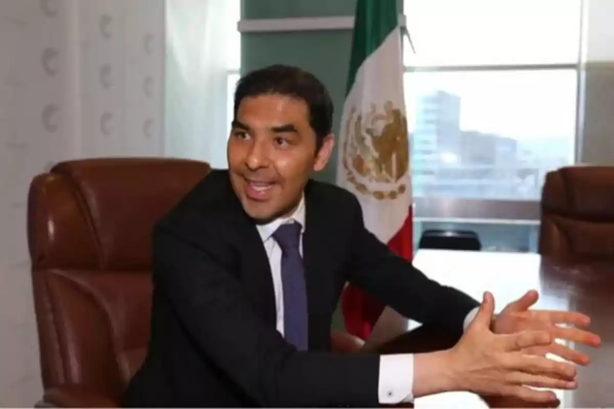 Un hombre en traje sentado en una oficina con una bandera de México al fondo.