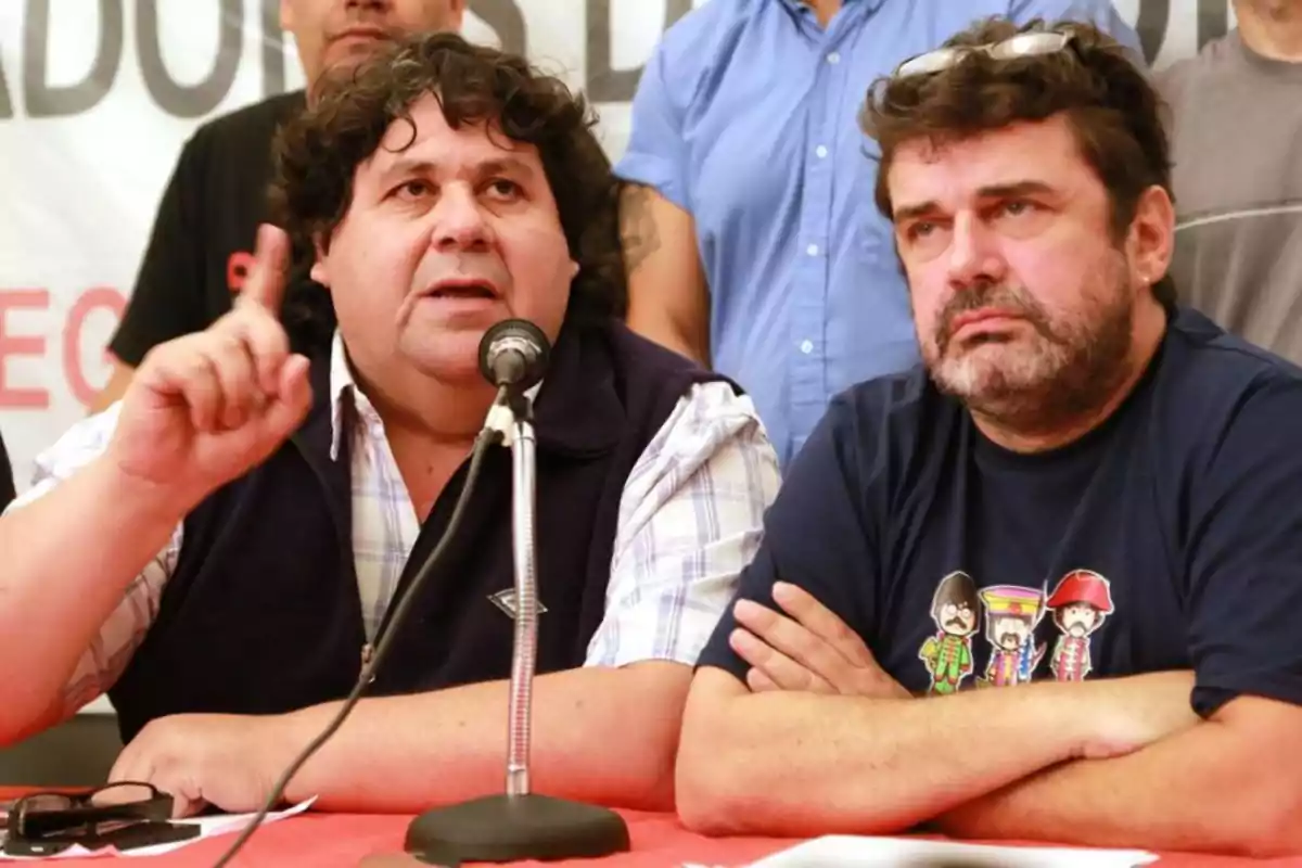 Dos hombres sentados en una mesa durante una conferencia de prensa uno de ellos habla por un micrófono mientras el otro escucha con los brazos cruzados.