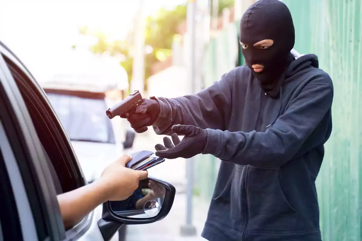 Person wearing a ski mask and holding a gun robbing a driver who is handing over his wallet through the car window