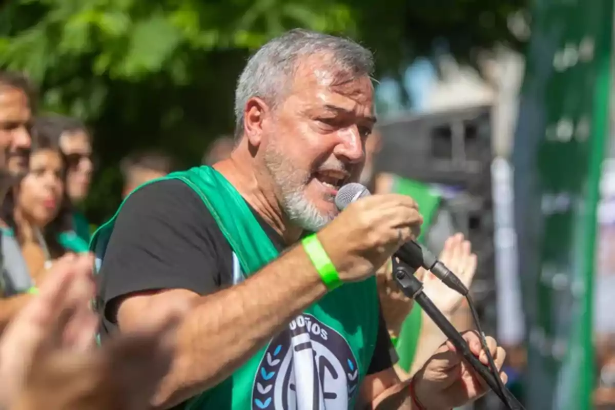 Gray-haired, bearded man speaking into a microphone during an outdoor protest with people in the background