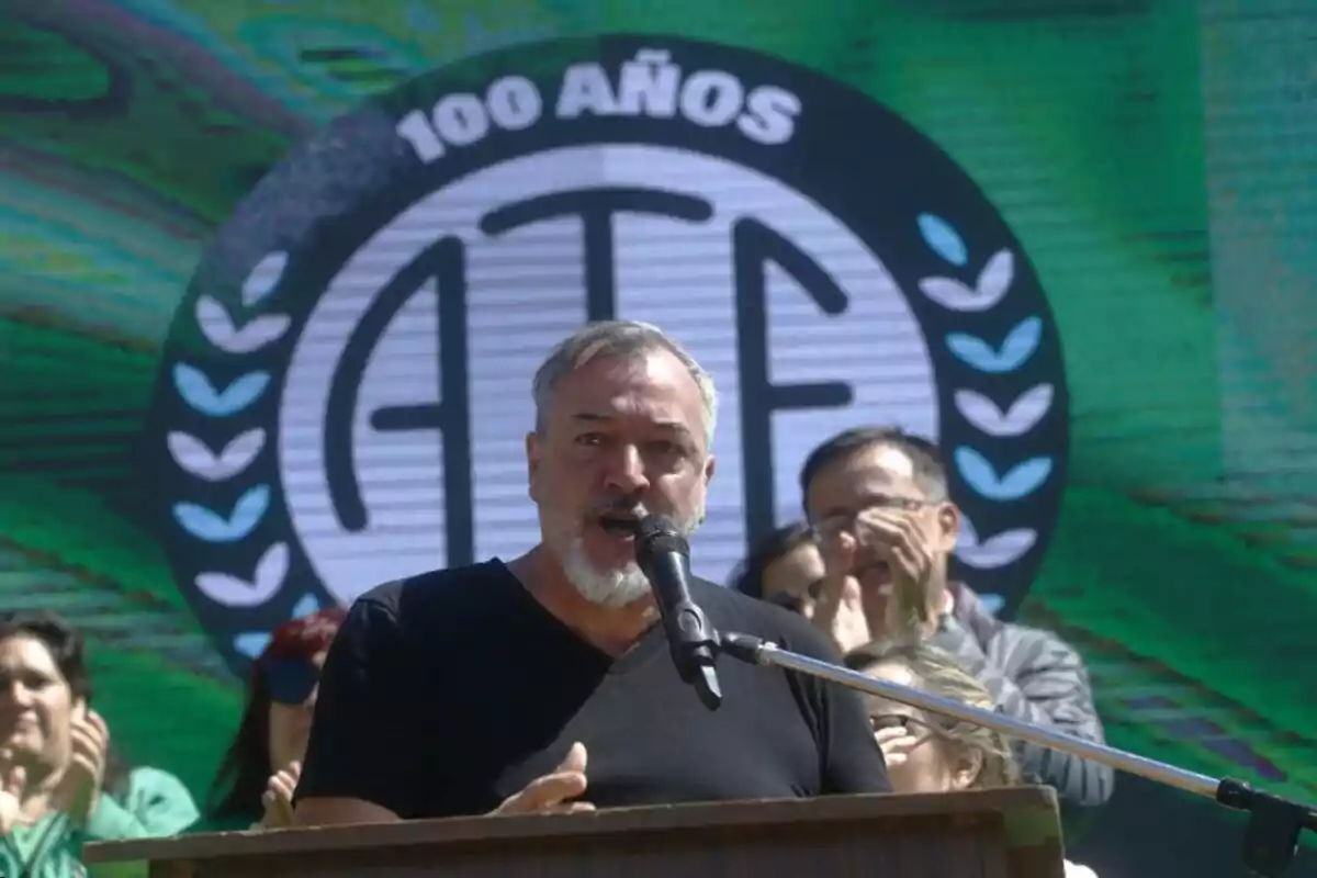 Un hombre hablando en un micrófono durante un evento de celebración del centenario de ATE, con un fondo verde y el logo de la organización.