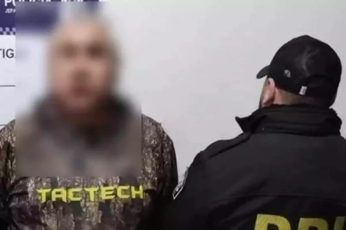 Un hombre con una sudadera de camuflaje y la palabra "TACTECH" está de pie frente a una pared, mientras que otro hombre con una gorra y una chaqueta con las letras "PDI" en la espalda lo observa.