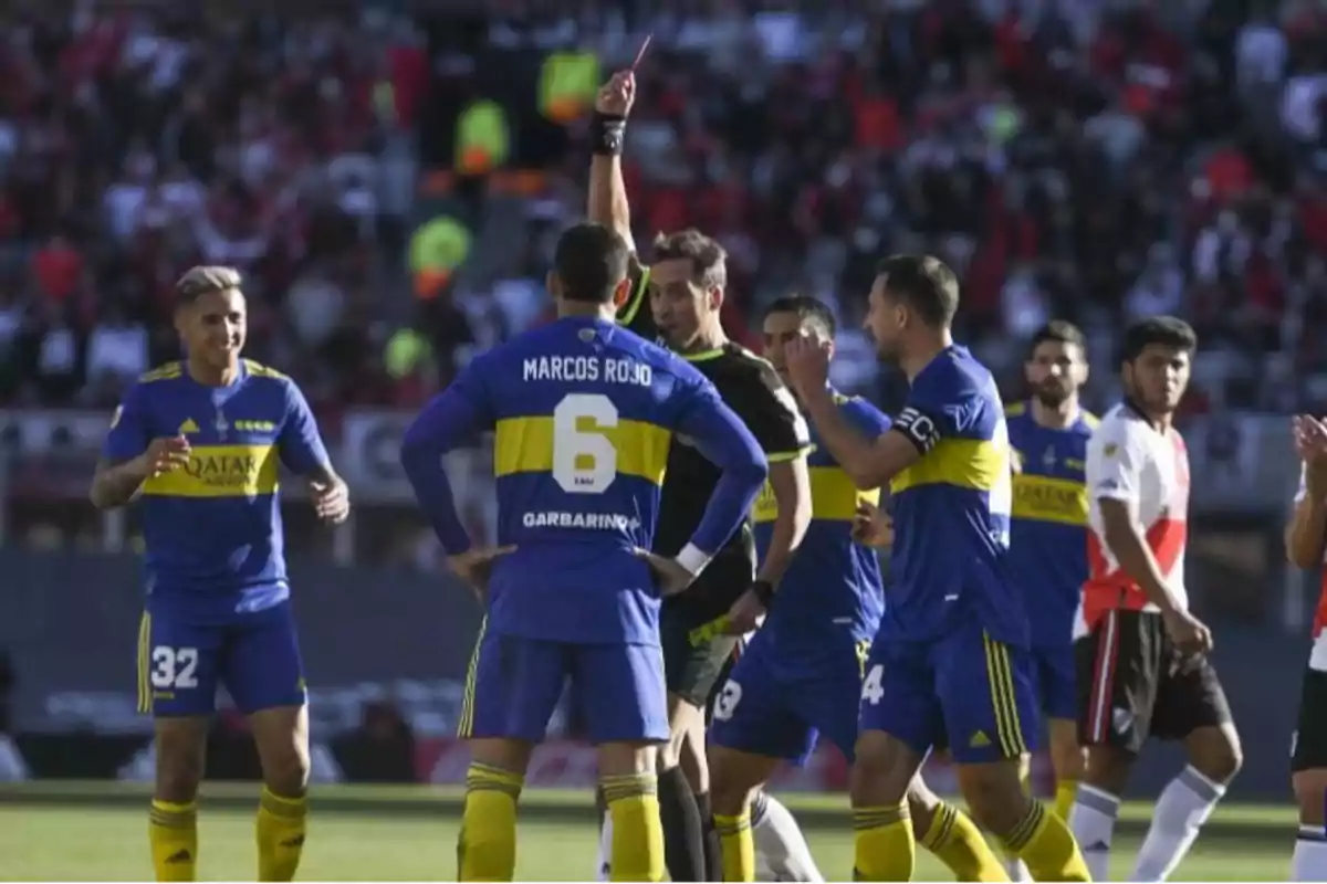 Jugadores de fútbol discuten con el árbitro mientras este muestra una tarjeta roja en un partido.