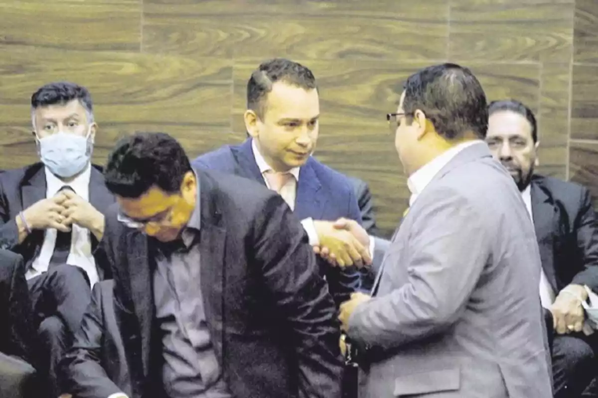 Un grupo de hombres en trajes formales se encuentra en una sala, dos de ellos se dan la mano mientras otros observan.