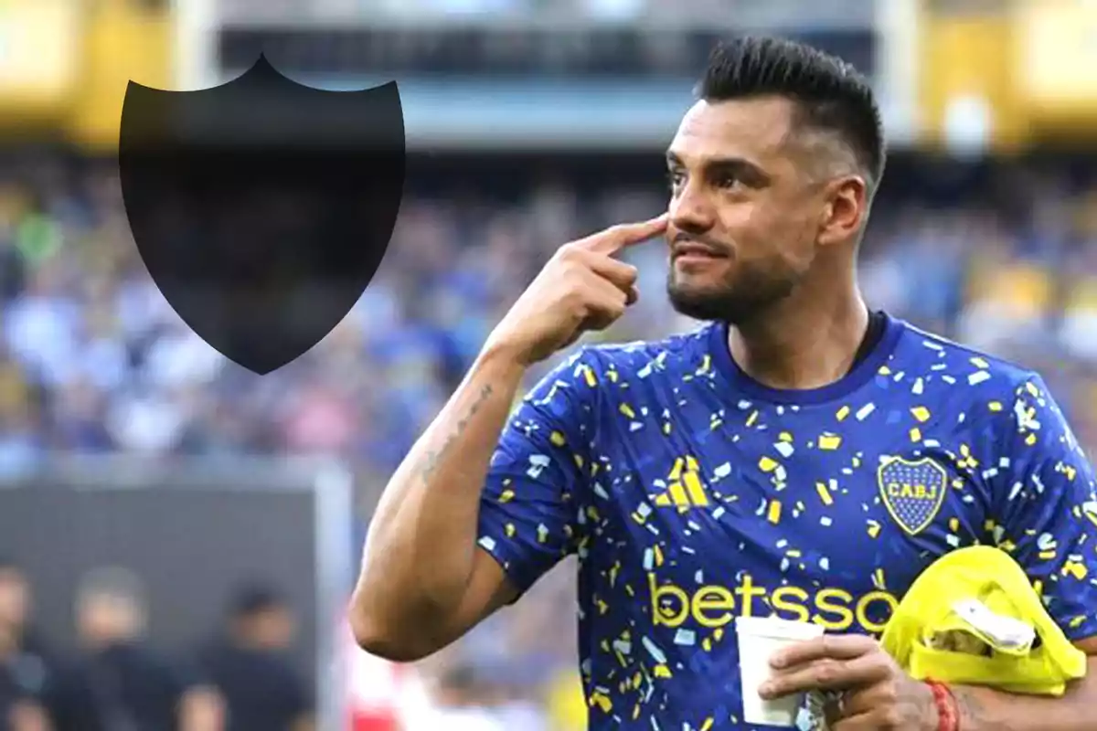 Un jugador de fútbol con la camiseta de Boca Juniors señala su rostro mientras sostiene un vaso y una toalla amarilla en un estadio lleno de espectadores.