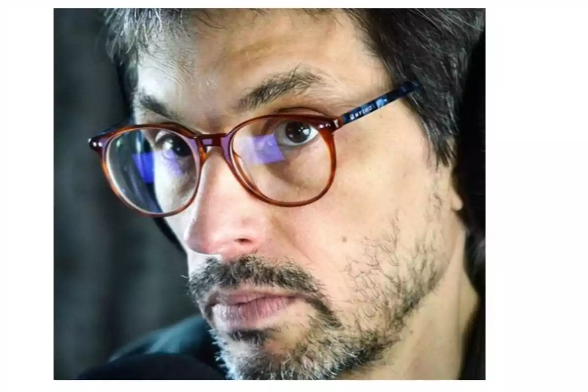 A man with a beard and brown-framed glasses looks forward with a serious expression. A man with a beard and brown-framed glasses looks forward with a serious expression.