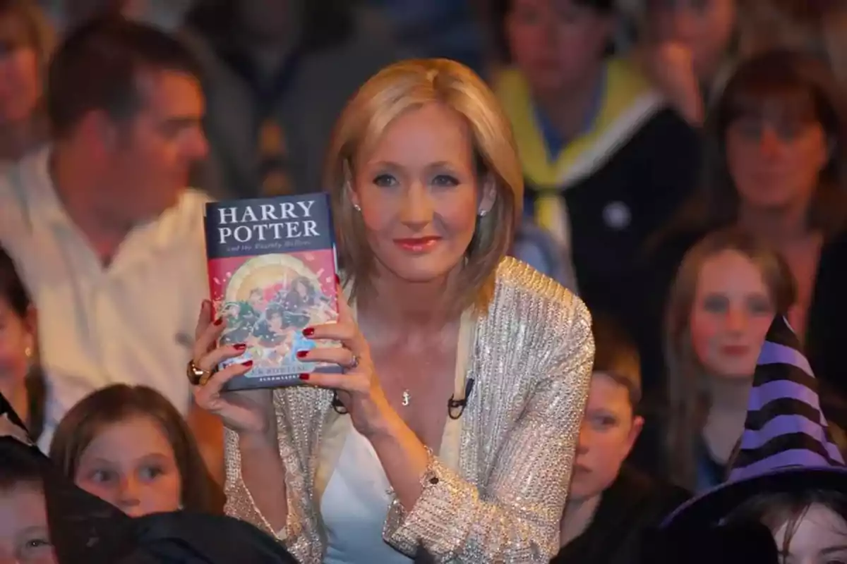 Mujer rubia con chaqueta brillante sostiene un libro de Harry Potter mientras está rodeada de personas en un evento Mujer rubia con chaqueta brillante sostiene un libro de Harry Potter mientras está rodeada de personas en un evento