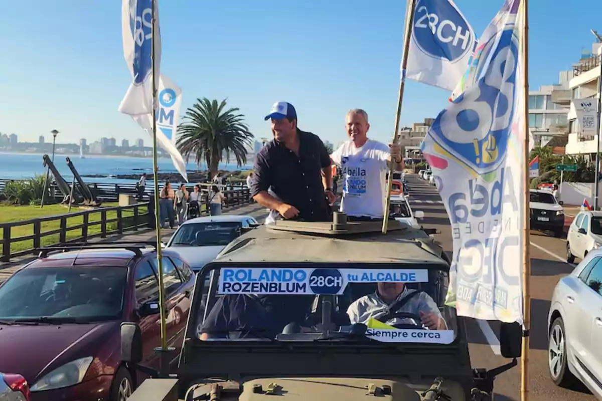 Un vehículo militar circula por una avenida costera con dos personas de pie en la parte superior, rodeado de banderas y carteles de campaña política, mientras varios autos lo siguen y un grupo de personas observa desde la acera.